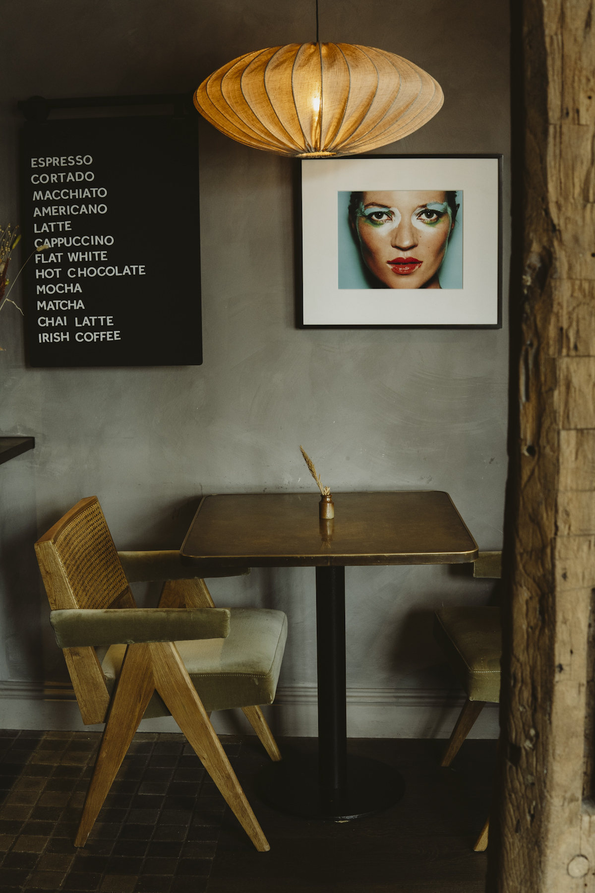 A cafe table and cane chair under a pleated pendant, beside a painted drinks list