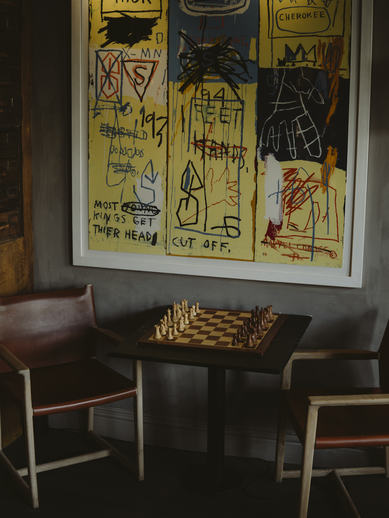 A chess set on a dark table below a large yellow artwork, flanked by leather chairs