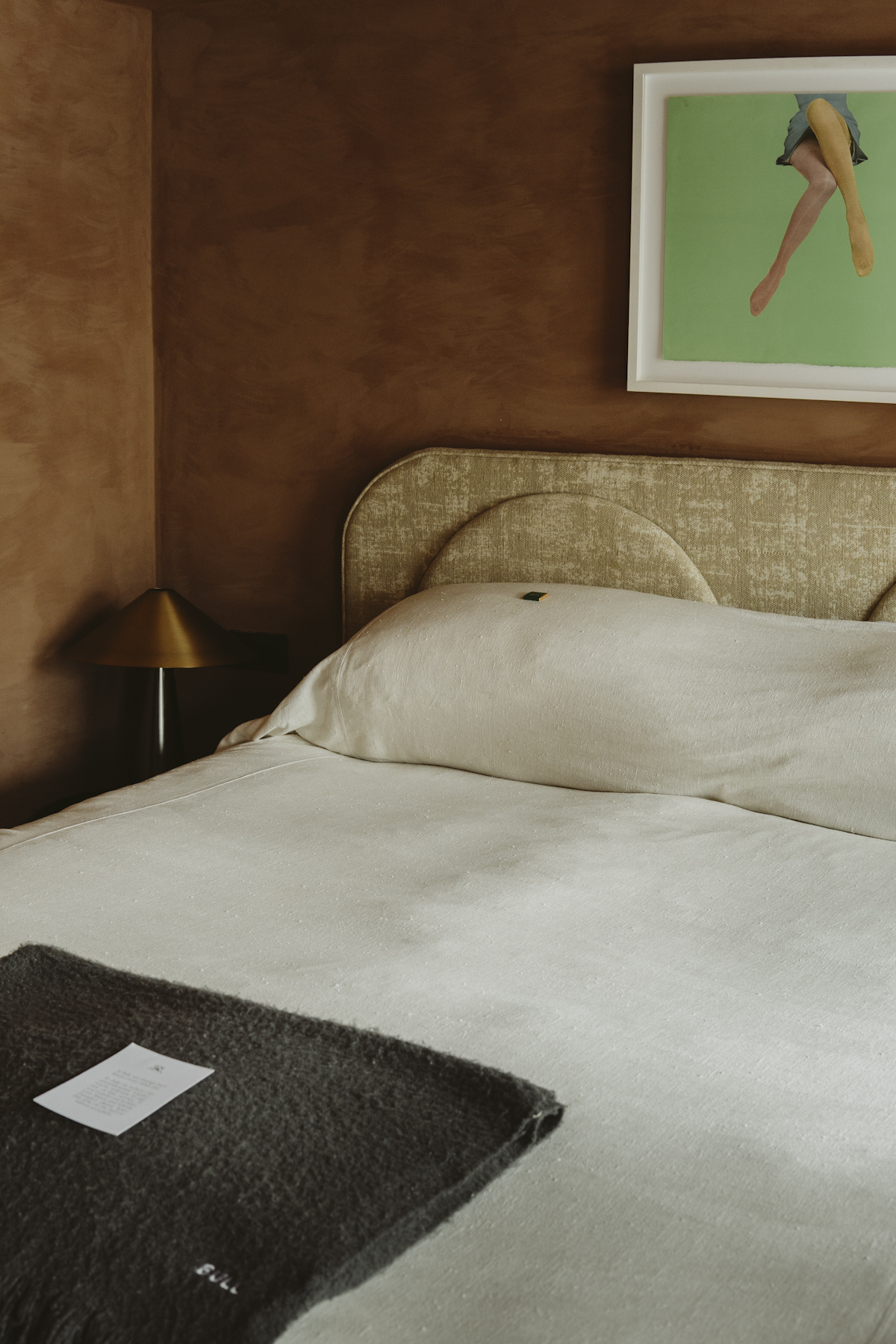 A made bed in cream linen with a grey throw, below a framed print on a plaster wall