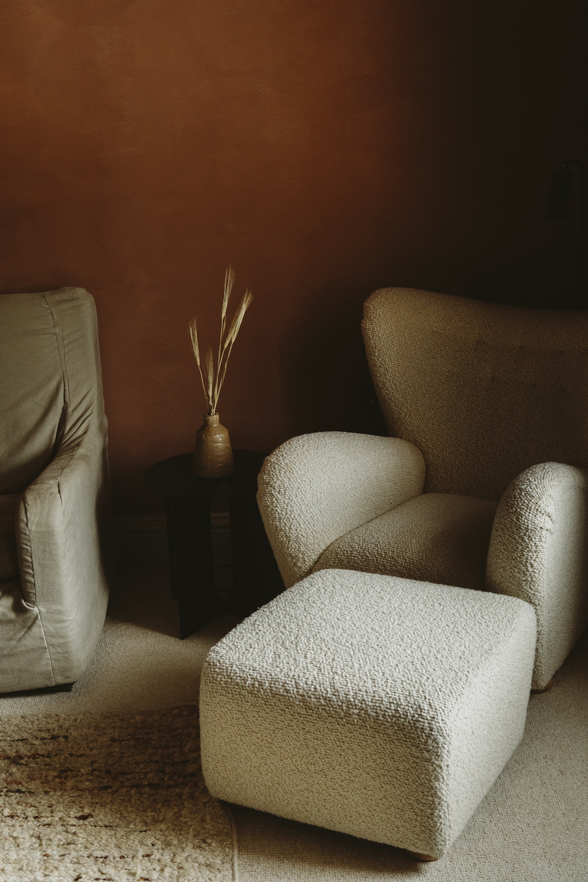 A boucle armchair and footstool against a deep brown wall